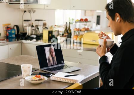 Young male freelancer having breakfast while doing video call with female colleague on laptop in kitchen Stock Photo