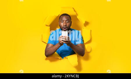 Shocked black man using cellphone, reading shocking message, posing in hole of torn paper on yellow background Stock Photo