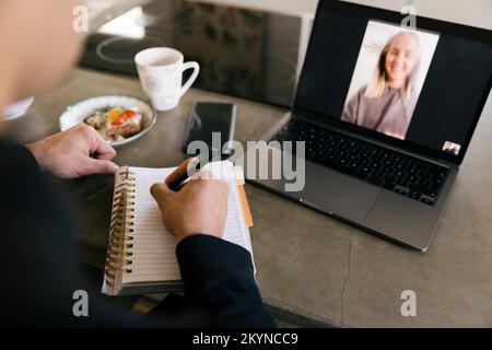 Hands of businessman writing in diary while doing video call with female colleague on laptop Stock Photo