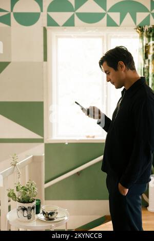 Side view of man with hand in pocket using smart phone while standing at home Stock Photo