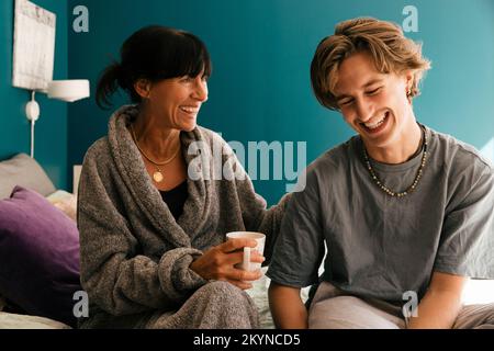 Woman holding mug while taking to happy son sitting on bed at home Stock Photo