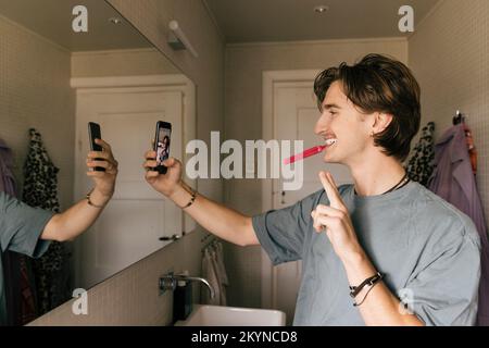Smiling man taking selfie through smart phone while brushing teeth in bathroom at home Stock Photo