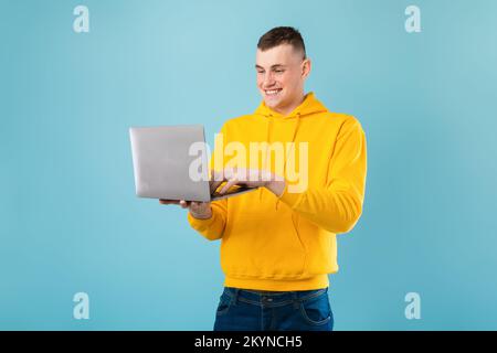 Happy handsome caucasian man enjoying his new modern laptop, typing on keyboard and smiling, blue background, copy space Stock Photo