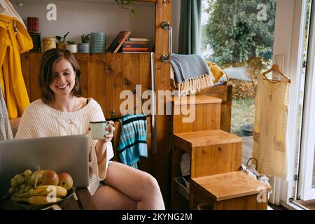 Smiling young woman using laptop holding coffee cup while sitting at home Stock Photo