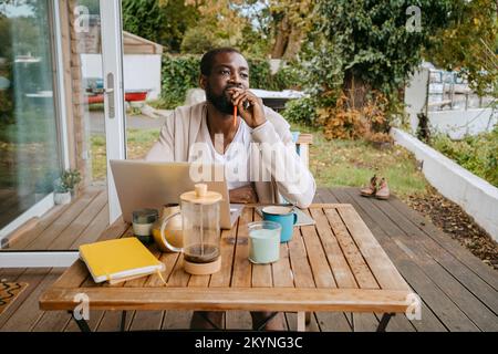 Thoughtful man with laptop sitting at table on porch Stock Photo