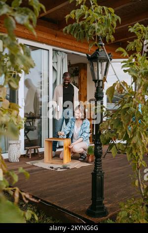 Happy couple helping each other while painting furniture on porch Stock Photo