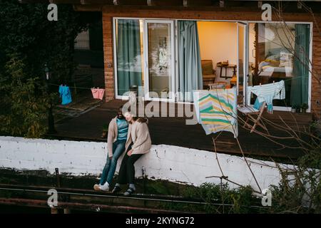 Multiracial couple enjoying with each other while sitting on retaining wall outside house Stock Photo