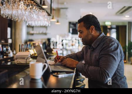 Businessman writing while sitting with laptop at bar counter Stock Photo