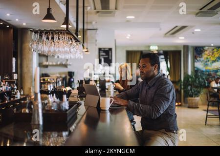 Male freelance worker using laptop while sitting at bar counter in restaurant Stock Photo