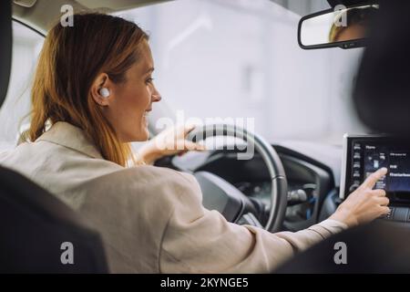 Smiling businesswoman wearing wireless in-ear headphones using dashboard control panel in car Stock Photo