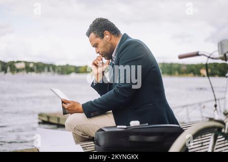 Side view of businessman looking at digital tablet while sitting on bench against bay Stock Photo