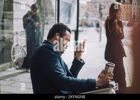 Side view of male professional sending voicemail while sitting against businesswoman talking on mobile phone at bus stop Stock Photo