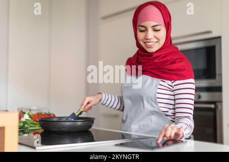 Beautiful muslim lady cooking at home, reading culinary blog Stock Photo