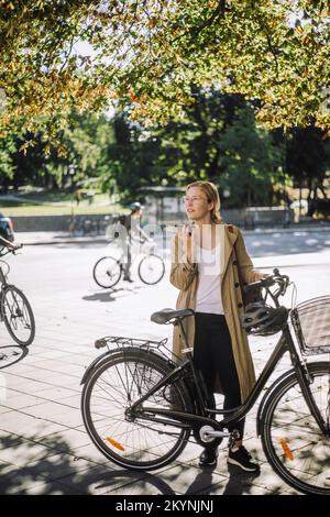 Young female commuter talking on speaker phone while standing with bicycle under tree Stock Photo
