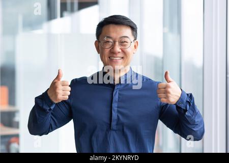 Portrait of successful asian man inside office near window, boss in glasses and shirt looking at camera and showing thumbs up. Stock Photo