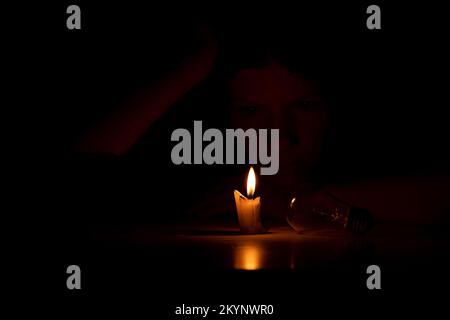 A girl in the dark sits with candles and a light bulb at a table in the dark at home, people without light at home Stock Photo