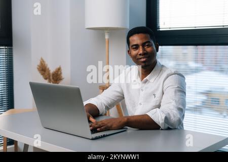 Portrait of positive African-American businessman using laptop and smiling looking at camera while enjoying work sitting at desk in modern office room Stock Photo