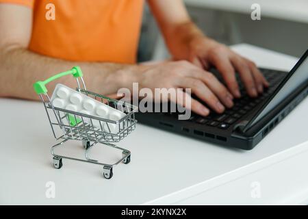 Small shopping trolley cart with blister pills on the table. Man using laptop for byuing tablets online. Purchase of medicines online. Stock Photo