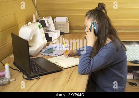 Woman fashion designer working on a laptop, making notes in a notebook, talking on the phone with a client. Small business. Stock Photo