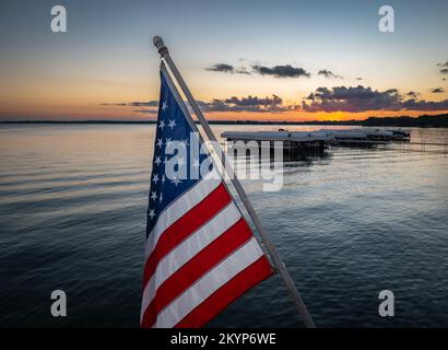 American flag on an angled pole with boats at a lake marina and water after sunset in the distance at dusk. Stock Photo