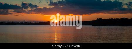 Serene sunset scene at a Minnesota lake with calm water and clouds and the sun setting behind trees on the distant shoreline. Stock Photo