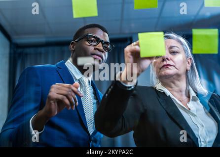 Serious mature businesswoman writing on sticky note in meeting room. Assistant business man planning and discussing strategy plan to boss. Stock Photo