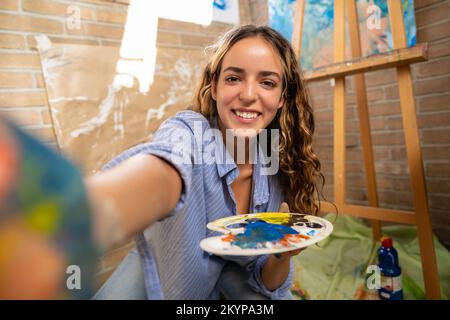 Young art student taking selfie while painting in her studio at home. Stock Photo