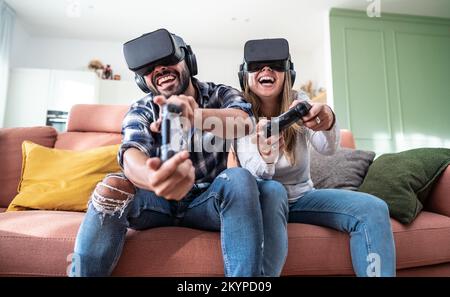 Happy couple playing video games with virtual reality glasses - Portrait of couple in VR glasses playing video game with joystick . Stock Photo