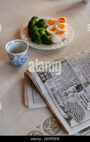 Boiled eggs, cut in half with bright orange yolks, and freshly blanched broccoli served at breakfast at home in Japan, with a newspaper and a cup of g Stock Photo