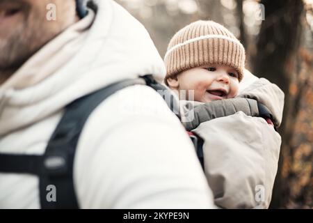 Sporty father carrying his infant son wearing winter jumpsuit and cap in backpack carrier hiking in autumn forest Stock Photo