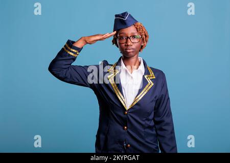 Portrait of female airline stewardess waving dressed in airline uniform. African american woman flight attendant looking confidently at camera in studio shot with blue background. Stock Photo