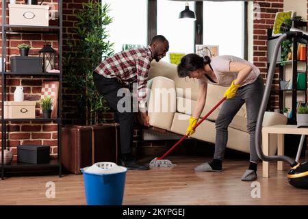 Multiracial couple doing spring cleaning at home, african american husband lifts heavy living room sofa for wife to clean underneath. Couple taking care of household chores together. Stock Photo
