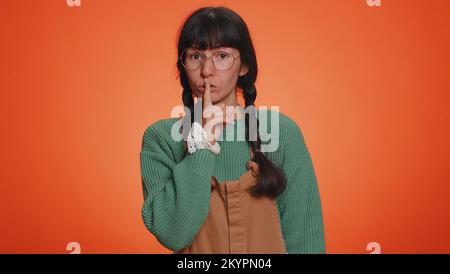 Shh be quiet please. Portrait of millennial adult woman 30 years old presses index finger to lips makes silence gesture sign do not tells secret. Young lovely girl posing on orange studio background Stock Photo