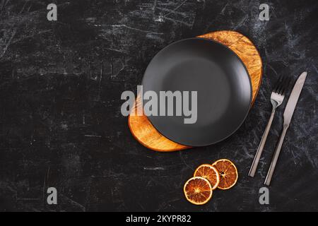 Black table setting with a plate on cutting board, fork and knife and orange slices. Top view, flat lay, copy space Stock Photo