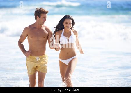 Spending some quality time together. A happy couple holding hands running in the waves. Stock Photo