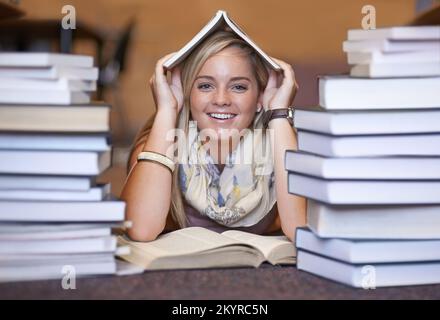 Knowledge resides between the covers. Portrait of a happy young college student sitting in the library with a book on her head. Stock Photo