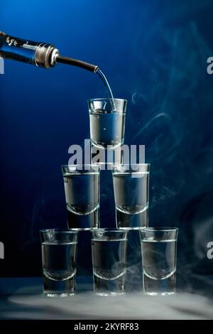 Vodka is poured from a bottle into a glass in smoke on dark blue background. Club drinks concept. Stock Photo