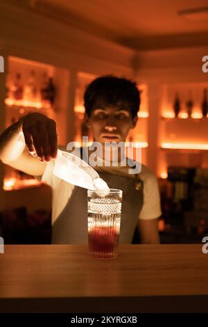 Professional bartender putting ice cubes to the cocktail glass with special spoon on the bar counter. Copy space Stock Photo