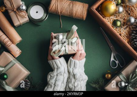 Woman s hands wrapping Christmas gift, close up. presents on olive background with decor elements and items, top view. Christmas or New year DIY packi Stock Photo