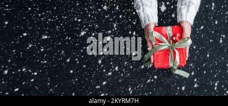 Female hands holding Christmas gift box on snowy black plaster background. Christmas background. Stock Photo