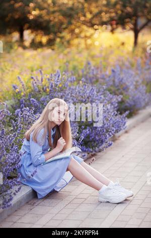 girl with pen writes in a notebook, sitting in the Park in the flowers outside. education and learning concept - happy student looking camera doing ho Stock Photo