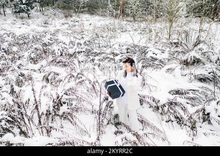 A stylish woman with a white suit with a New Year's gift in her hands in a winter forest. A girl in nature in a snowy forest with a gift box Stock Photo