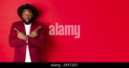 Christmas shopping and people concept. Indecisive Black man choosing between two variants, pointing fingers sideways and staring confused camera, red Stock Photo
