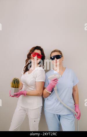 Two cosmetologists in protective glasses are holding a laser depilation device and a cactus in their hands Stock Photo