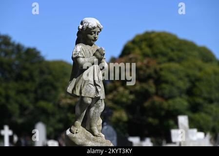 Old, worn, and weathered stone sculpture of a robed child looking down mournfully, hands clasped together in prayer, in a cemetery Stock Photo