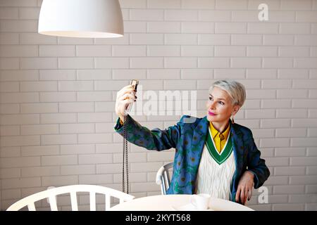 Mature short gray hair woman holds smartphone, taking selfie, sitting at table in coffee shop with cup against white wall background. Female dressed i Stock Photo