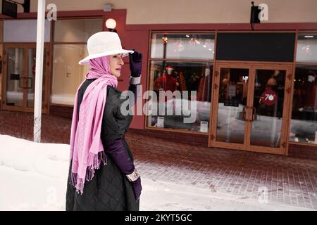 Elegant mature woman in stylish outfit shopping near discount clothing store at winter street with shops, wearing headscarf, fedora hat, side view Stock Photo