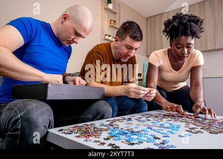 Friends playing with jigsaw puzzle at home, on a white wooden table. Putting things together and solving problems. Fun and diversity in friendship. Stock Photo