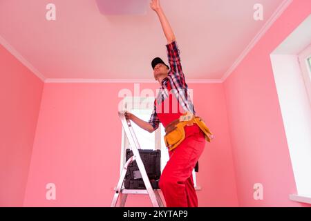 Electrician changing light bulb. Young man in overall work uniform standing on ladder in residential building, office, school or hospital and screwing Stock Photo