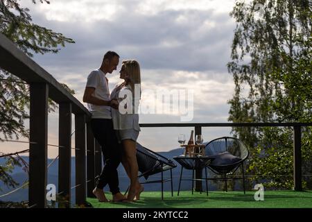 Horizontal sideview shot of a young couple in summer outfit enjoying the mountain view from terrace. Copy space. Stock Photo